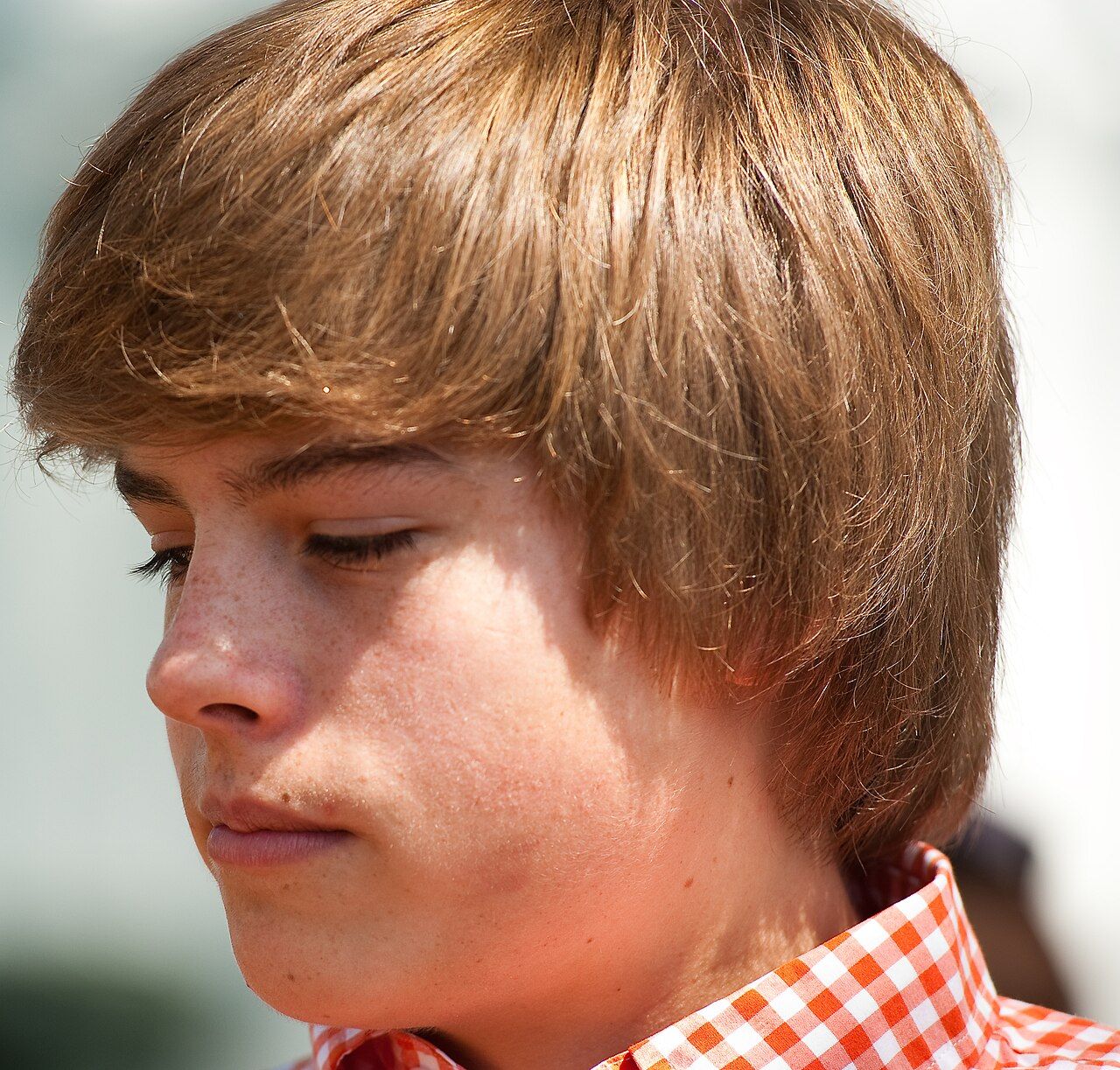 A young man with shaggy brown hair looks down, wearing a red checked shirt