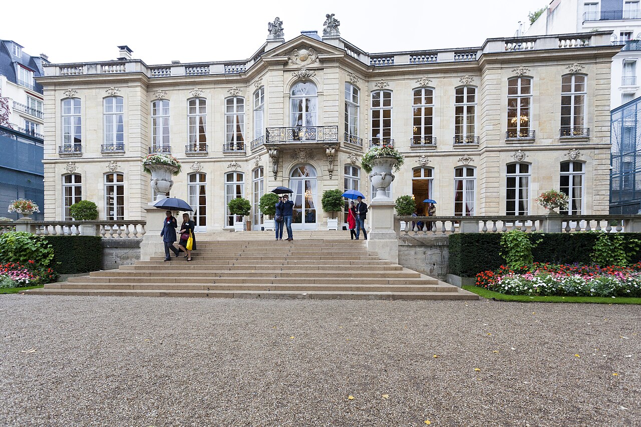 A pale stone mansion with tall windows and wide steps facing a gravel courtyard