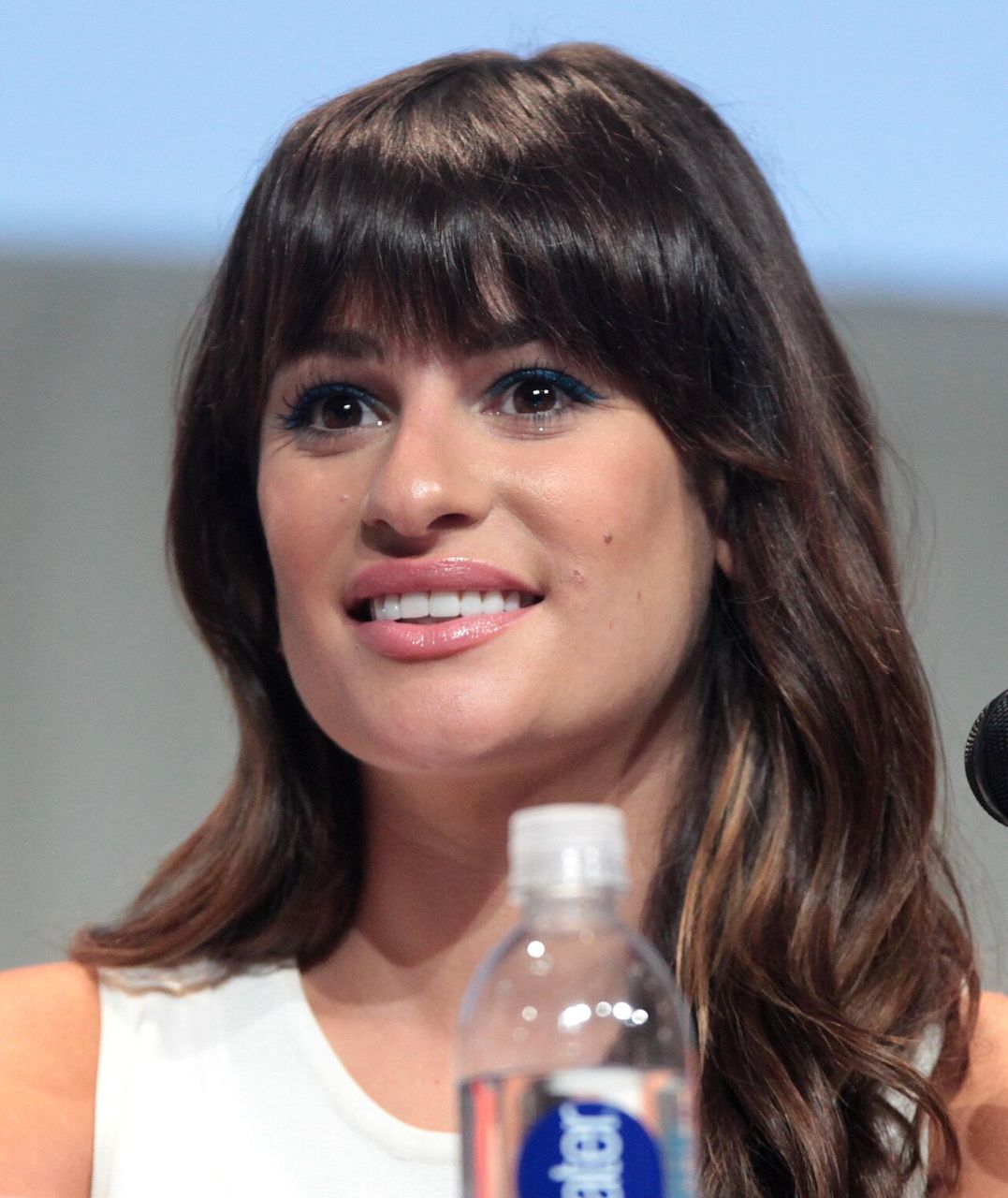Lea Michele, with long dark hair, smiles at a panel with a water bottle in front of her