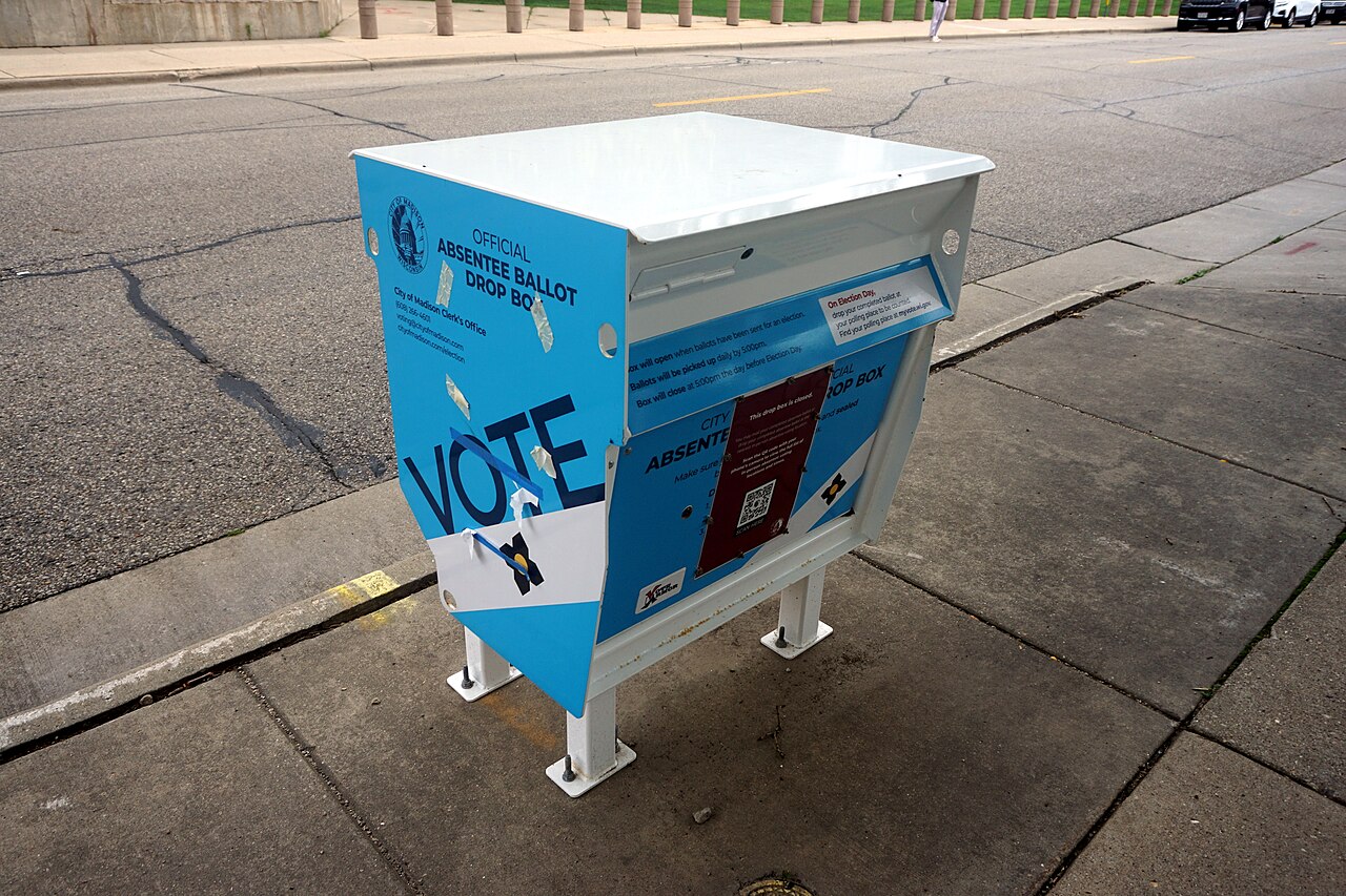 A blue metal ballot drop box with a VOTE sign stands on a concrete sidewalk