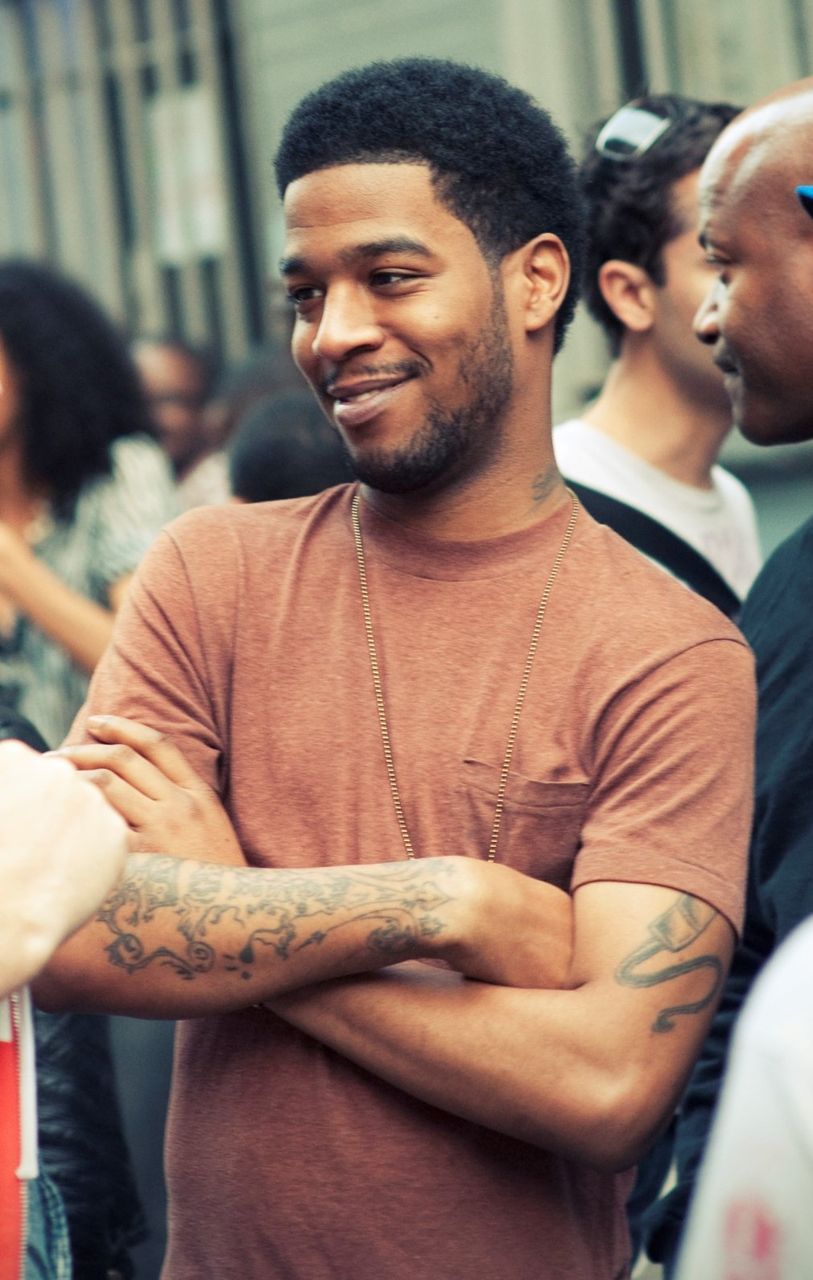 Kid Cudi in an orange shirt smiles with his arms crossed outdoors