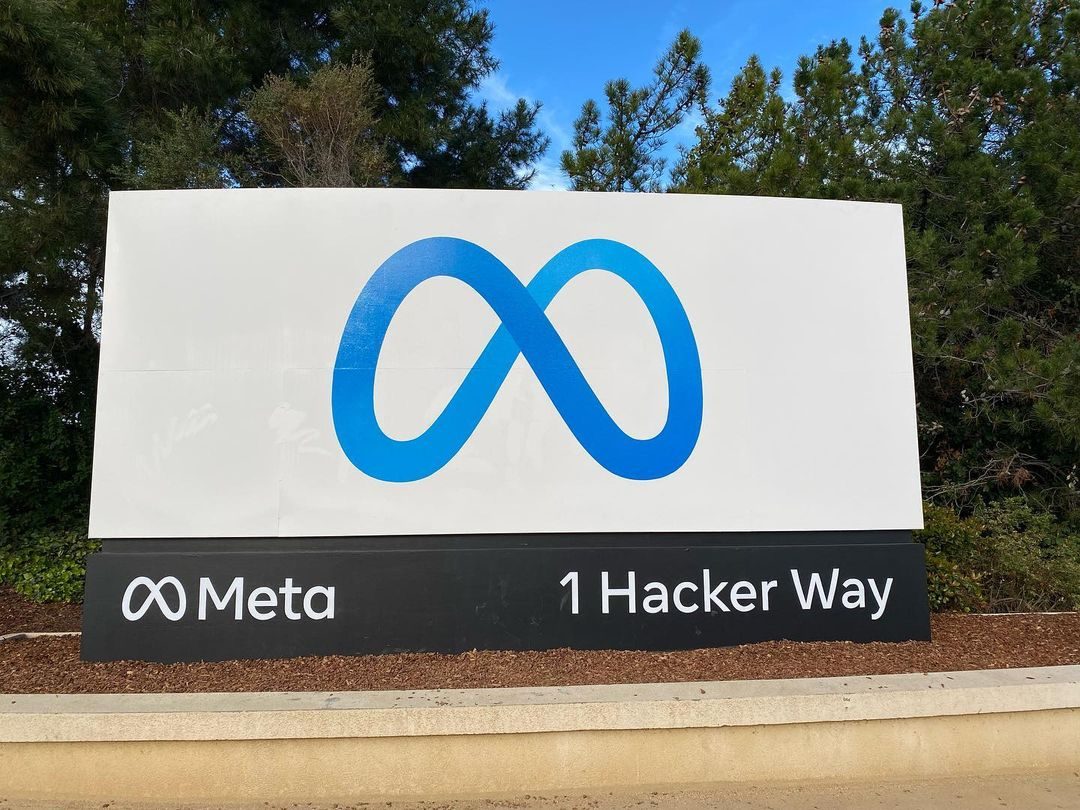 A white sign with Meta's blue infinity logo and the address 1 Hacker Way, with trees behind it