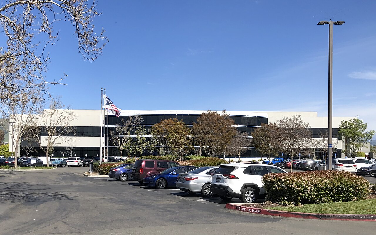 A low white office building with a flag out front behind a parking lot lined with cars and trees