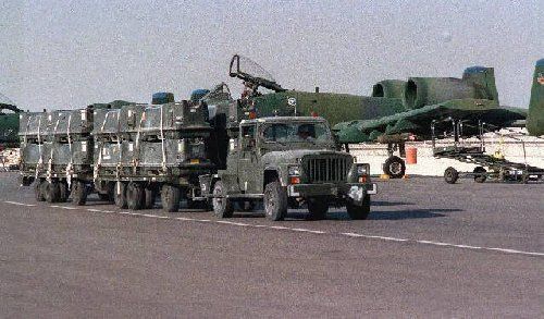 A truck hauls equipment along an airfield in front of a row of parked military jets