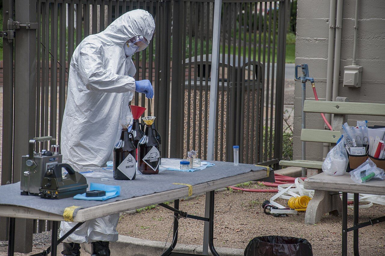 A worker in a white protective suit, goggles and blue gloves pours chemicals into labeled bottles on a table