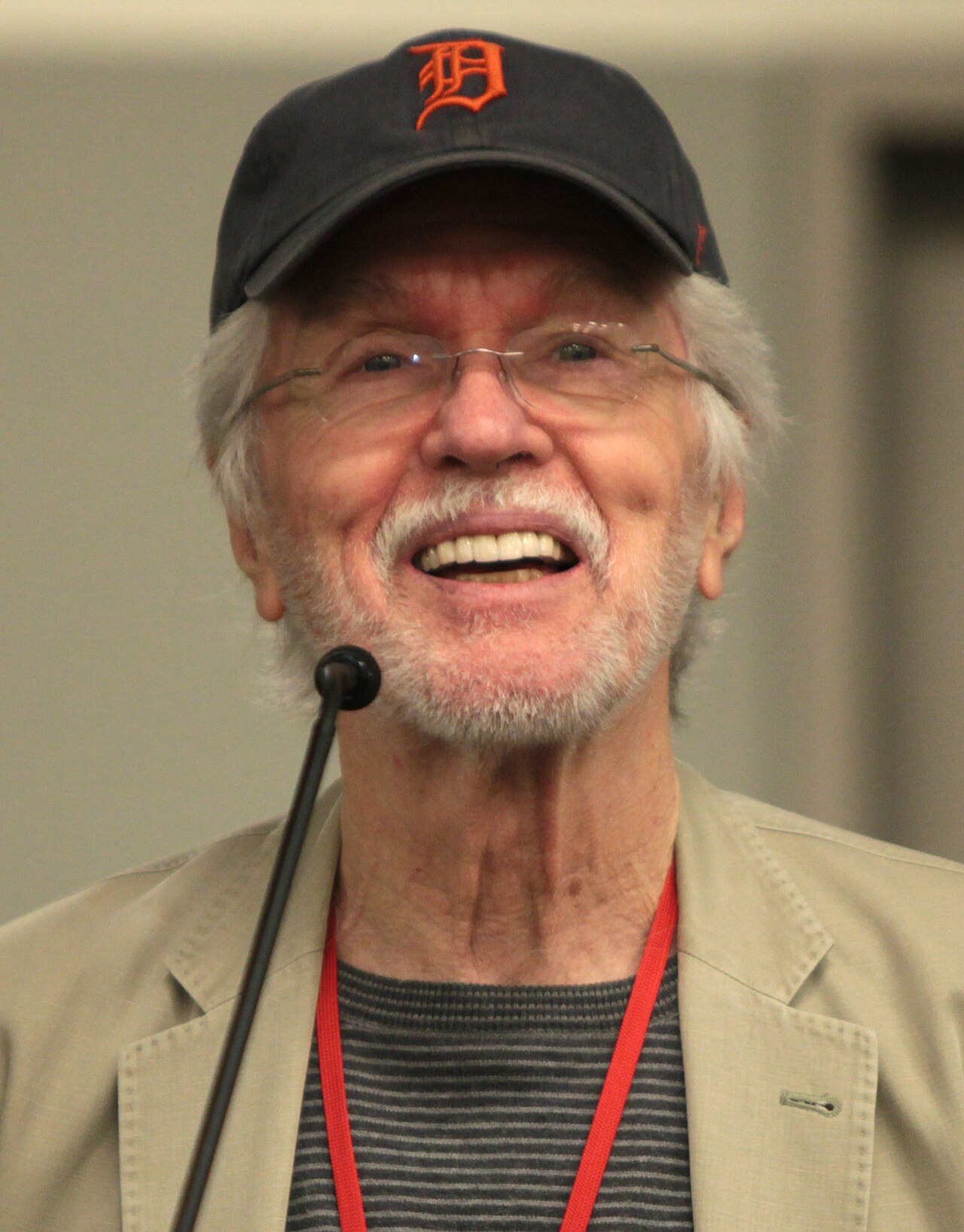 An older man with a white beard, glasses and a baseball cap smiles while speaking into a microphone