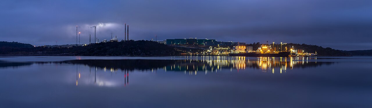 Fog drifts over the towers of an oil refinery at dusk beside a fjord