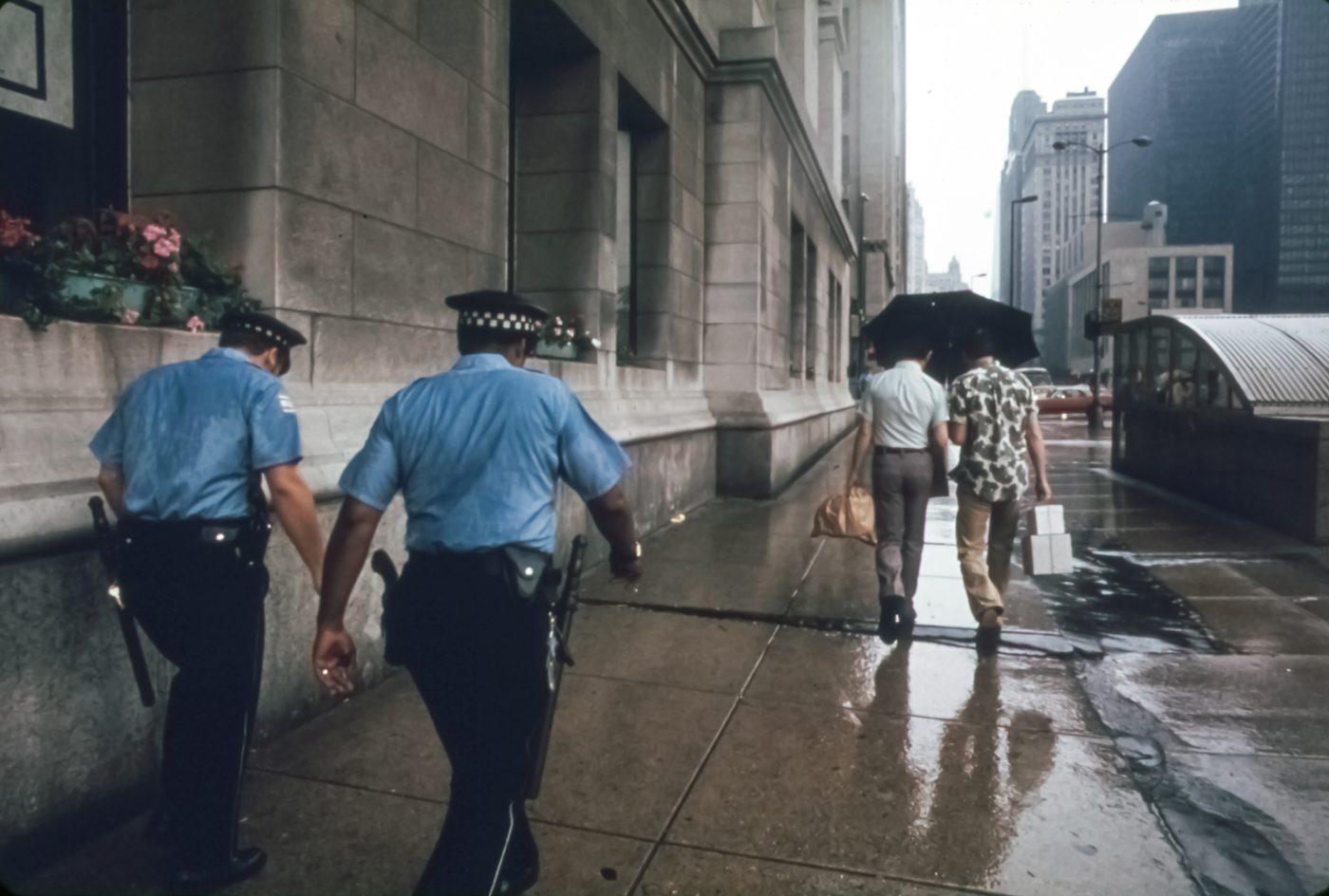 People with umbrellas and two police officers walk along a wet sidewalk on North Michigan Avenue in Chicago on a rainy day