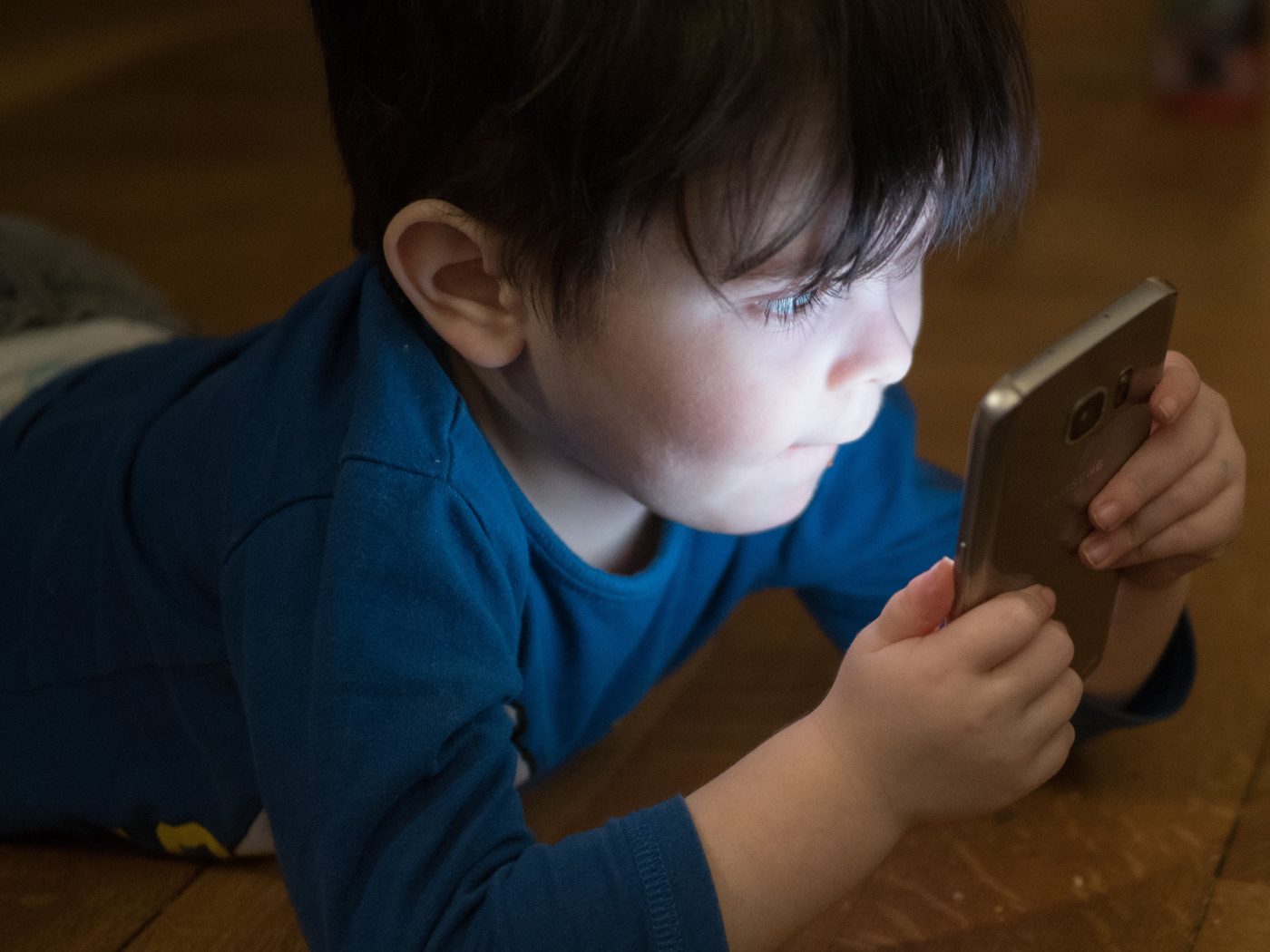A boy lying on a carpet looking at a glowing smartphone