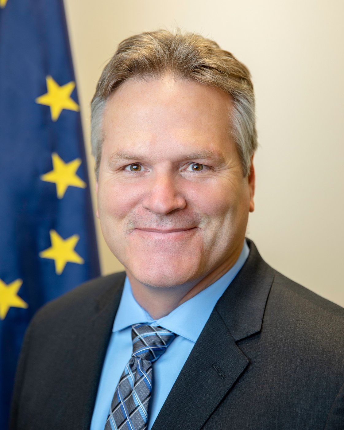 Alaska Governor Mike Dunleavy smiles in a dark suit in front of a flag