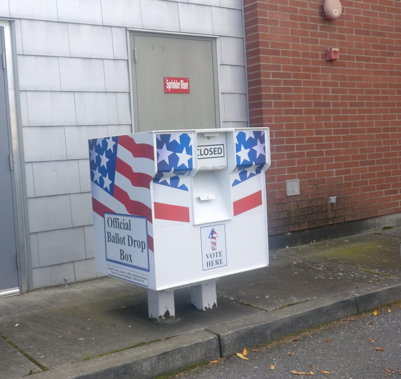 A red, white and blue ballot drop box stands on a sidewalk beside a building
