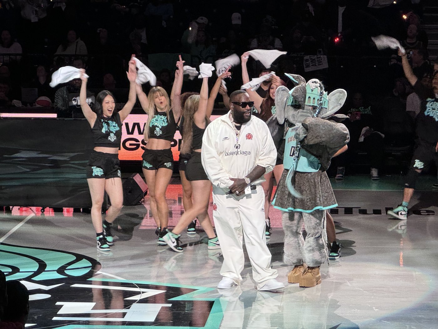 Rapper Rick Ross in a white outfit stands on a basketball court with dancers
