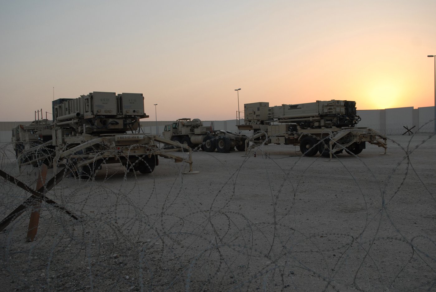 Patriot missile launchers sit in a desert field behind barbed wire at sunset